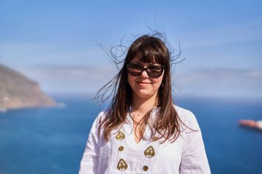 Rüzgarlı saçları olan, güneş gözlüğü takan ve Tenerife 'de okyanus kenarında beyaz işlemeli elbisesi olan gülümseyen bir kadın.