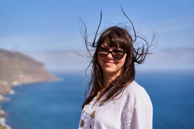 Rüzgarlı saçları olan, güneş gözlüğü takan ve Tenerife 'de okyanus kenarında beyaz işlemeli elbisesi olan gülümseyen bir kadın.