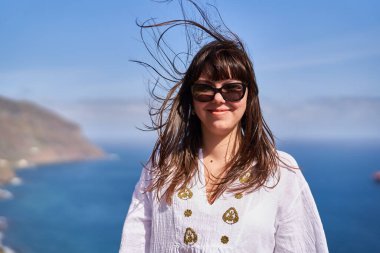 Rüzgarlı saçları olan, güneş gözlüğü takan ve Tenerife 'de okyanus kenarında beyaz işlemeli elbisesi olan gülümseyen bir kadın.