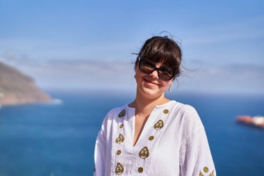 Güneş gözlüğü takan gülümseyen bir kadın ve Tenerife 'nin arka planında uzak bir gemi olan beyaz işlemeli bir elbise.