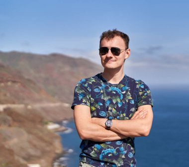 Güneş gözlüğü takan kendinden emin bir adam ve Tenerife 'de okyanusun kenarında kolları çapraz duran çiçek desenli bir gömlek.