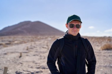 Güneş gözlüğü, kep ve sırt çantası takan gülümseyen bir adam Tenerif 'te volkanik bir tepeyle kuru bir yürüyüş yolunda duruyordu.