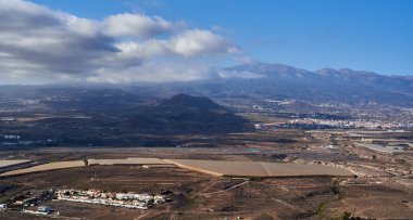 Tenerife manzarası, kayan tepeler, tarlalar ve vadide yuvalanmış küçük kasabalar, arka planda dağlar ve yukarıda dağınık bulutlar.