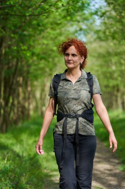 Güneşli bir günde yemyeşil bir ormanda gezen sırt çantalı orta yaşlı bir kadın.