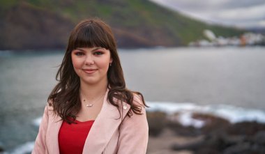 Pembe ceketli ve kırmızı bluzlu güzel bir esmer, okyanusun kenarında duruyor yumuşak doğal ışıkta kendine güvenen bir gülümsemeyle.