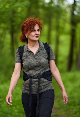 Güneşli bir günde yemyeşil bir ormanda gezen sırt çantalı orta yaşlı bir kadın.