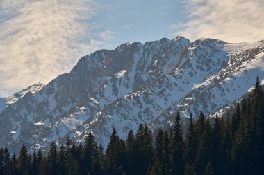 Karla kaplı engebeli dağ zirveleri yoğun bir yeşil ormanın üzerinde parçalı bulutlu bir gökyüzünün altında yükseliyor.