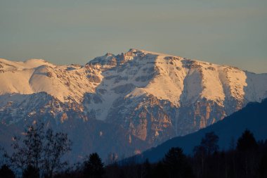 Karla kaplı engebeli dağ zirvesi kayalık uçurumlar ve sıcak akşam ışığında yemyeşil ormanlar.