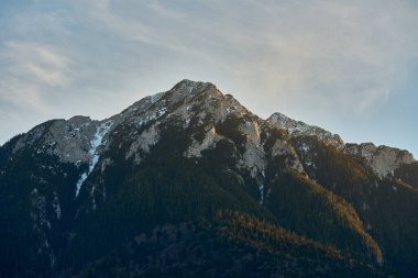 Karla kaplı engebeli dağ zirvesi kayalık uçurumlar ve sıcak akşam ışığında yemyeşil ormanlar.