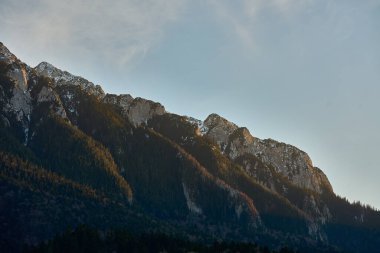 Karla kaplı engebeli dağ zirvesi kayalık uçurumlar ve sıcak akşam ışığında yemyeşil ormanlar.