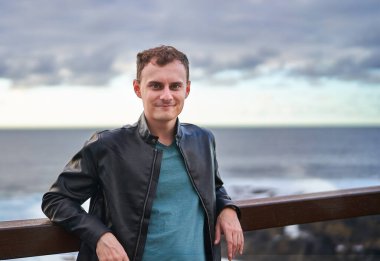 Siyah deri ceket giyen genç bir gezgin Tenerife 'de kayalık bir kıyıya vuran dalgalarla okyanusun kenarında duruyor.