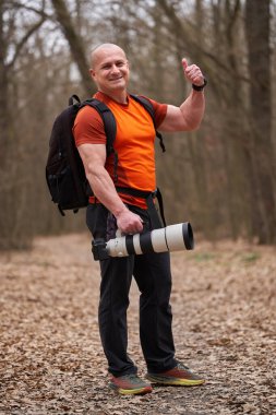 Gülümseyen adam kamerayı telefonla tutuyor ve ormanda baş parmağını kaldırıyor.