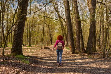 Kızıl saçlı kadın sırt çantalı yalnız başına orman yolunda yürüyor. Arkadan görülüyor.