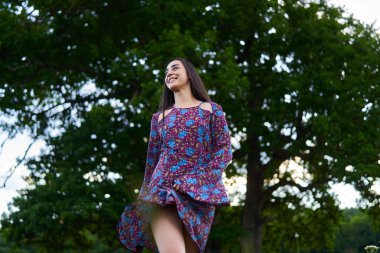 Çiçekli elbiseli neşeli genç bir kadın. Gülümsüyor ve arka planda uzun ağaçlarla yeşil bir çayırda yürüyor.