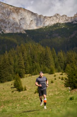 Çimenli dağ yolunda koşan adam açık mavi gökyüzünün altında arkasında kayalık tepeler var.