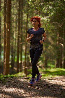 Güneşli orman yolunda koşan bir kadın spor giyim ve koşu ayakkabıları giyiyor.