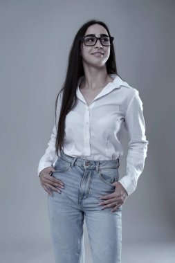 Kot pantolonlu, beyaz gömlekli, gözlüklü, hafif bir gülümsemeyle poz veren, gri arka planda soyutlanmış, kendinden emin genç bir kadın.
