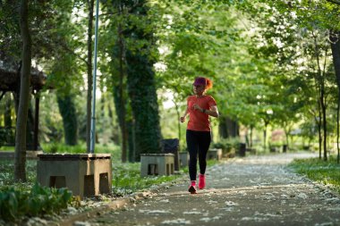 Güneşli bir yaz gününde yapraklı park yolunda koşan bir kadın.