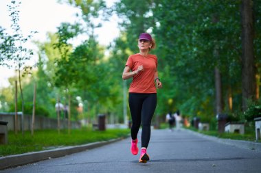 Güneşli bir yaz gününde yapraklı park yolunda koşan bir kadın.