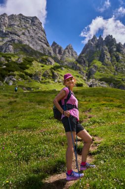 Güneşli bir dağ yamacında, kayalık arka planda yaban çiçekleri arasında yokuş yukarı yürüyen bir kadın.