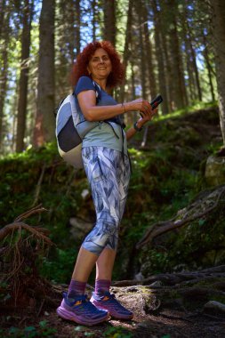 Sırt çantası ve telefon elinde benekli ışığın altında orman yolunda gülümseyen kadın.