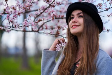 Çiçek açan bahar ağacının altında pembe çiçeklerle gülümseyen ve siyah şapkalı genç bir kadın.