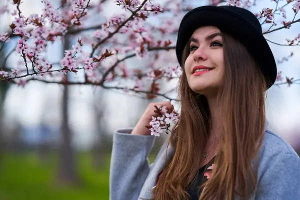 Çiçek açan bahar ağacının altında pembe çiçeklerle gülümseyen ve siyah şapkalı genç bir kadın.