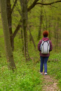 Beyaz sırt çantalı bir kadın dar patika boyunca canlı bahar ormanlarında tek başına yürüyüş yapıyor.