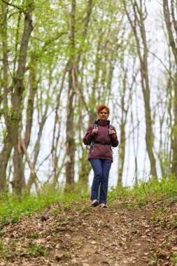 Kızıl saçlı, olgun bir kadın. Sırt çantasıyla geziyor. Dar bir patikada bereketli bahar ormanlarında.