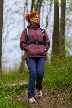 Kızıl saçlı, olgun bir kadın. Sırt çantasıyla geziyor. Dar bir patikada bereketli bahar ormanlarında.