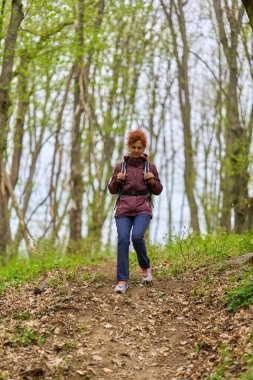 Kızıl saçlı, olgun bir kadın. Sırt çantasıyla geziyor. Dar bir patikada bereketli bahar ormanlarında.