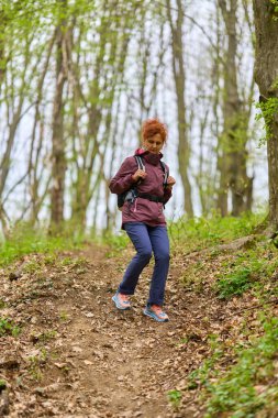 Kızıl saçlı, olgun bir kadın. Sırt çantasıyla geziyor. Dar bir patikada bereketli bahar ormanlarında.
