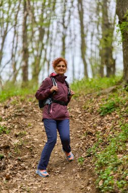 Kızıl saçlı, olgun bir kadın. Sırt çantasıyla geziyor. Dar bir patikada bereketli bahar ormanlarında.