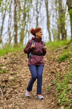 Kızıl saçlı, olgun bir kadın. Sırt çantasıyla geziyor. Dar bir patikada bereketli bahar ormanlarında.