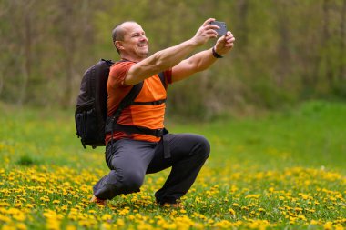 Çiçekli bir bahar çayırında çömelmiş bir adam akıllı telefonuyla selfie çekiyor ve baş parmağını kaldırıyor.