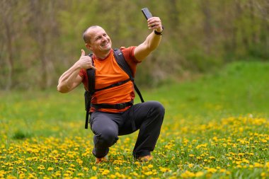 Çiçekli bir bahar çayırında çömelmiş bir adam akıllı telefonuyla selfie çekiyor ve baş parmağını kaldırıyor.