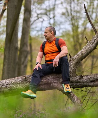 Turuncu gömlekli erkek yürüyüşçü bahar ormanı ve vahşi bitki örtüsüyle çevrili devrilmiş bir ağaçta dinleniyor.