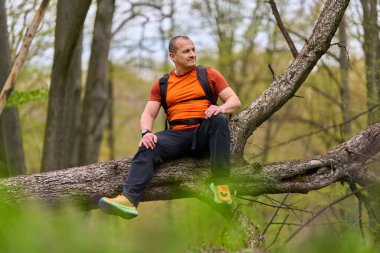 Turuncu gömlekli erkek yürüyüşçü bahar ormanı ve vahşi bitki örtüsüyle çevrili devrilmiş bir ağaçta dinleniyor.