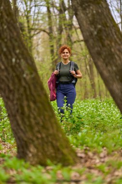 Kıvırcık dişi sırt çantalı gezgin bir bahar günü sakin bir orman patikasında tek başına yürüyüş yapıyor.