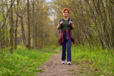 Kıvırcık dişi sırt çantalı gezgin bir bahar günü sakin bir orman patikasında tek başına yürüyüş yapıyor.