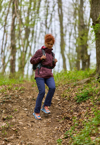 Redhead mature woman with backpack hiking alone through lush spring forest on a narrow path