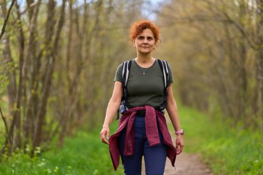 Orta yaşlı, kızıl kıvırcık saçlı bir kadın yeşil orman yolunda güvenle yürüyor.