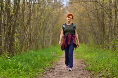 Orta yaşlı, kızıl kıvırcık saçlı bir kadın yeşil orman yolunda güvenle yürüyor.