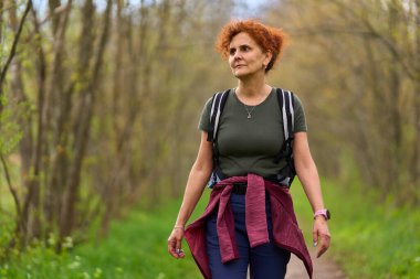 Orta yaşlı, kızıl kıvırcık saçlı bir kadın yeşil orman yolunda güvenle yürüyor.