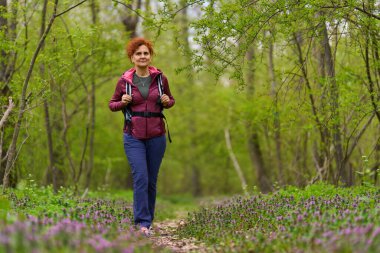 Orta yaşlı, kızıl kıvırcık saçlı bir kadın yeşil orman yolunda güvenle yürüyor.