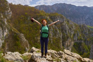 Neşeli kadın yürüyüşçü kollarını kaldırmış kayalık bir dağın tepesinde, yamaçlarda ve ilkbaharda orman yamaçlarında dikiliyor.