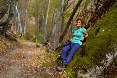 Bir dişi yürüyüşçü, orman patikasının yanındaki yosunlu bir ağaç gövdesine oturur. Elinde bir kamera ve gülümser.
