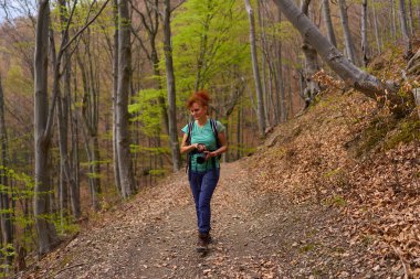 Bir kadın elinde kamerayla orman yolunda yürüyor ve baharın başında sırt çantası takıyor.