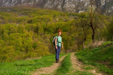 Sırt çantalı bir kadın yemyeşil bir dağda çimenli bir patikada dururken gülümsüyor.