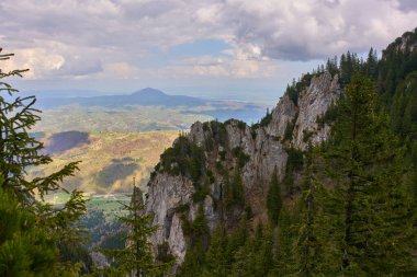 Parçalı bulutlu bir gökyüzünün altında geniş kırsal bir vadiye bakan çam ormanlı dramatik dağ uçurumları